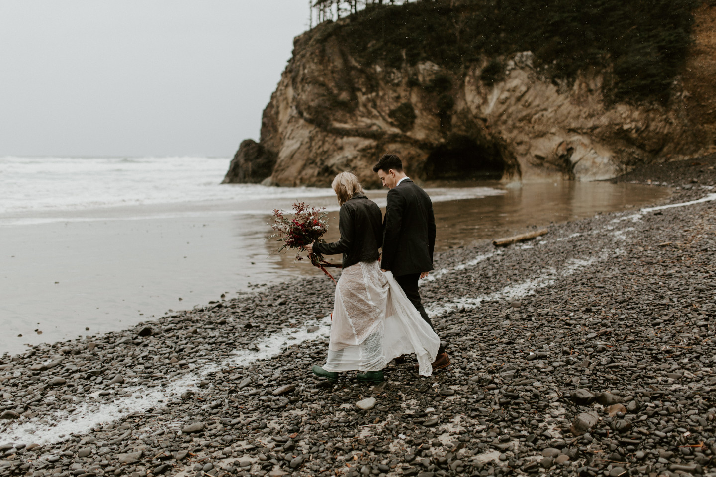 Hannah and Grant walk along the rocky shore of Cannon Beach during their rainy elopement in Oregon. Wedding photography in Portland Oregon by Sienna Plus Josh.