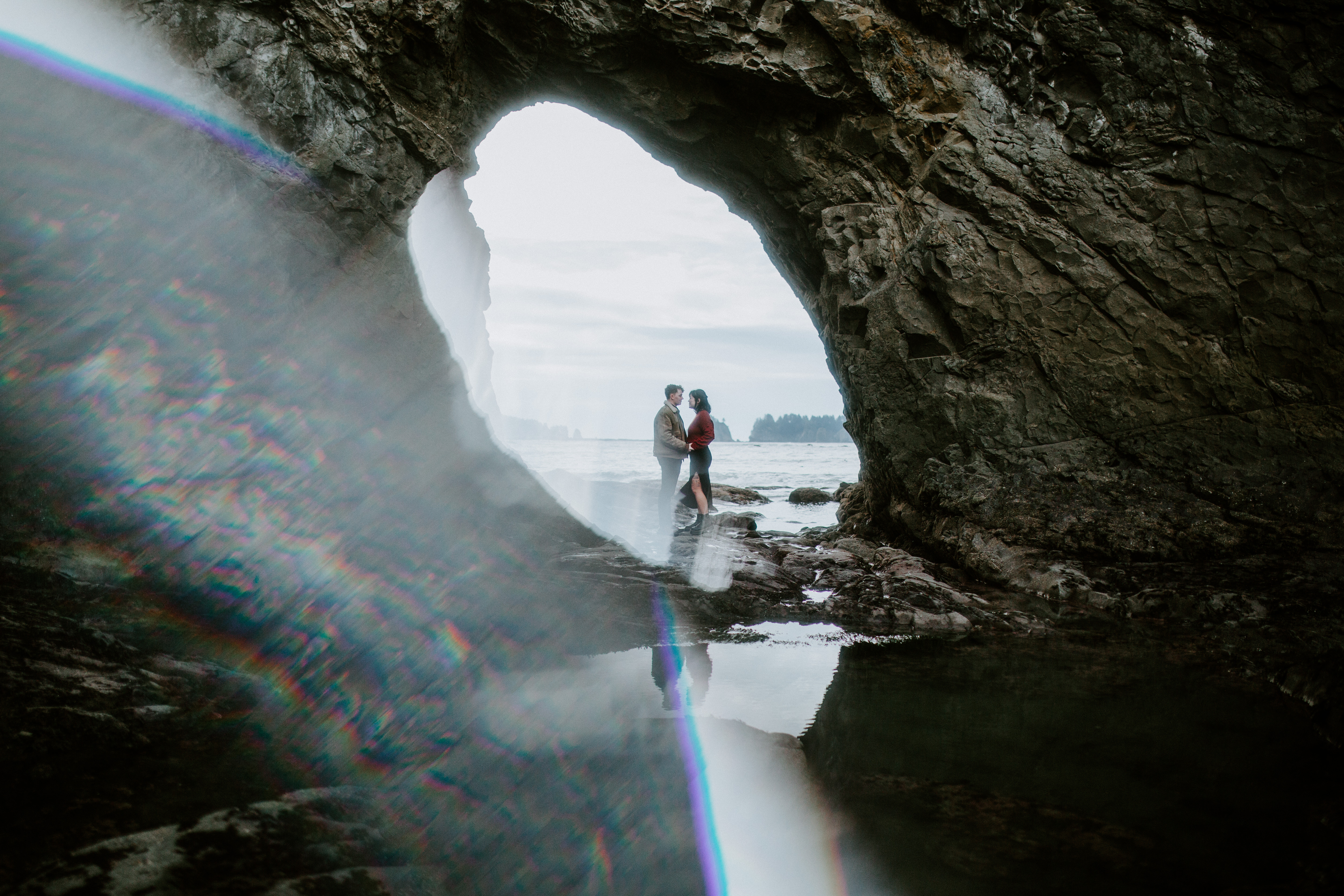 Bethany and Brett stand on the sand of Rialto Beach, under a rock arch.
